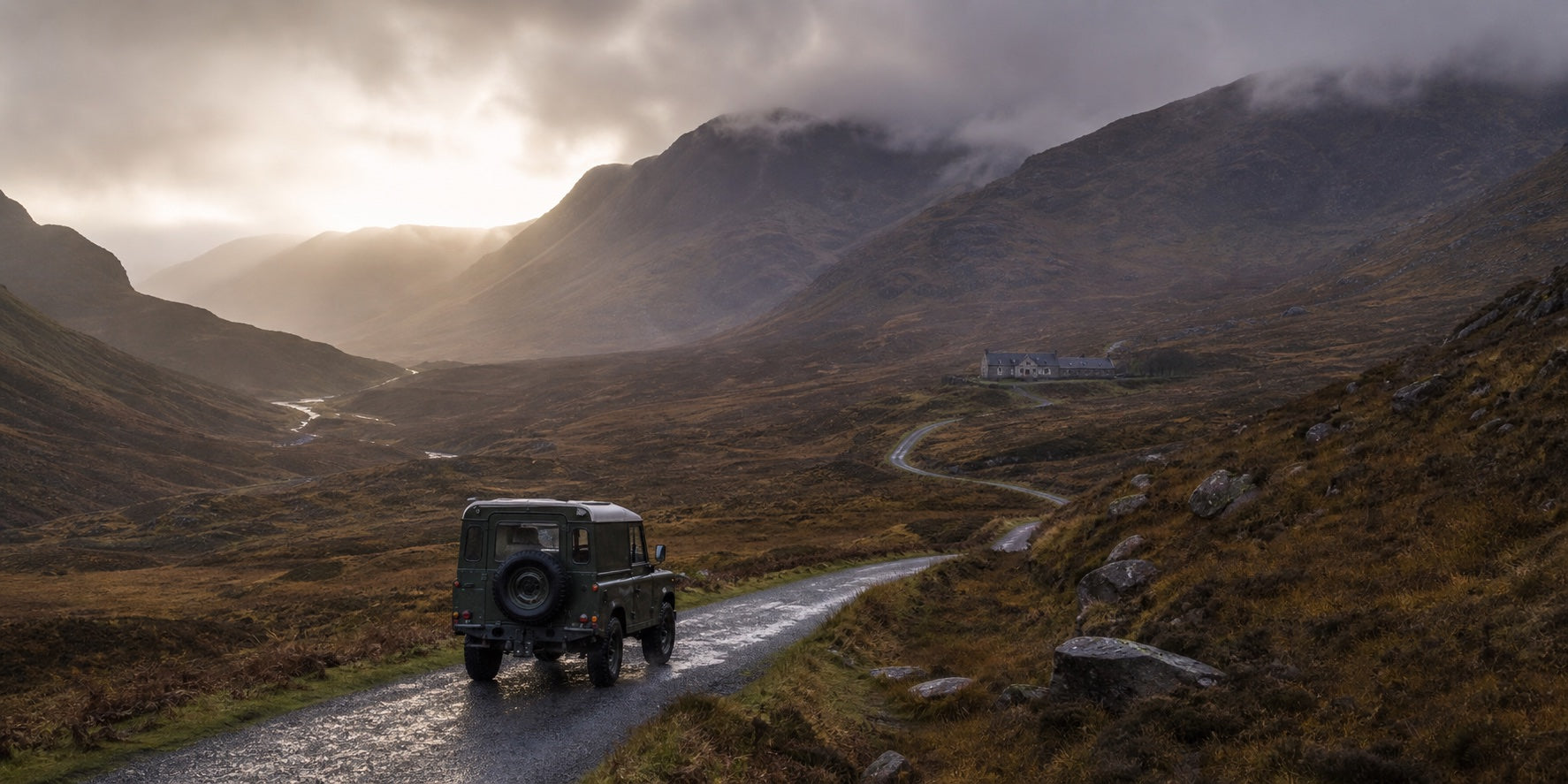 A dark olive four-wheel drive travelling through the Scottish Highlands towards a remote stone retreat at dawn